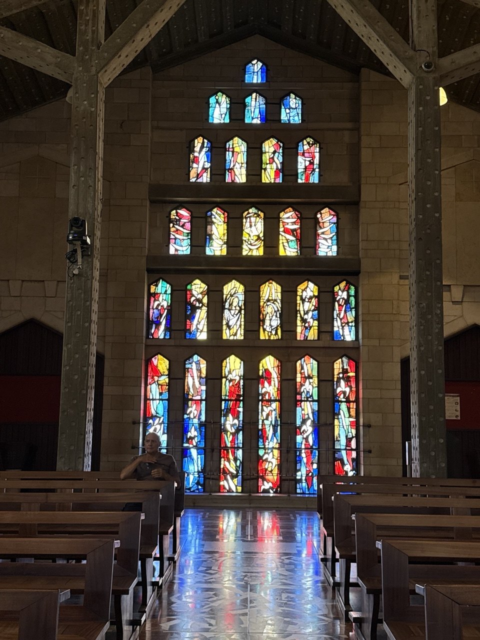 Nave gereja Maria, dinding kaca patri setinggi nave. Warna-warninya jatuh dan menggenang di lantai marmer.