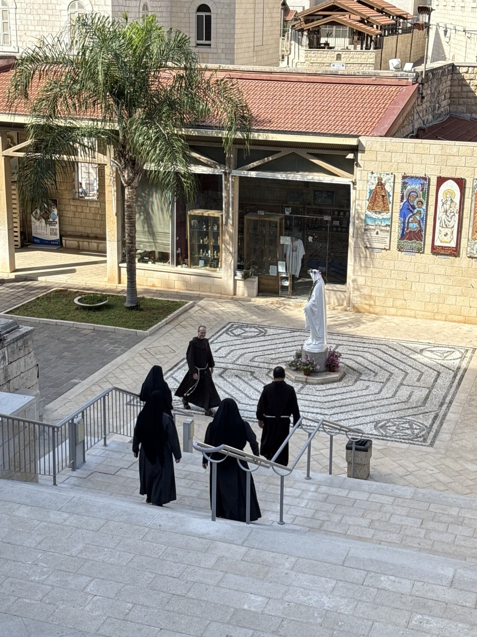 Halaman bawah basilika, suster-suster Fransiskan menuruni tangga menuju patung Maria putih di atas lantai mosaik.